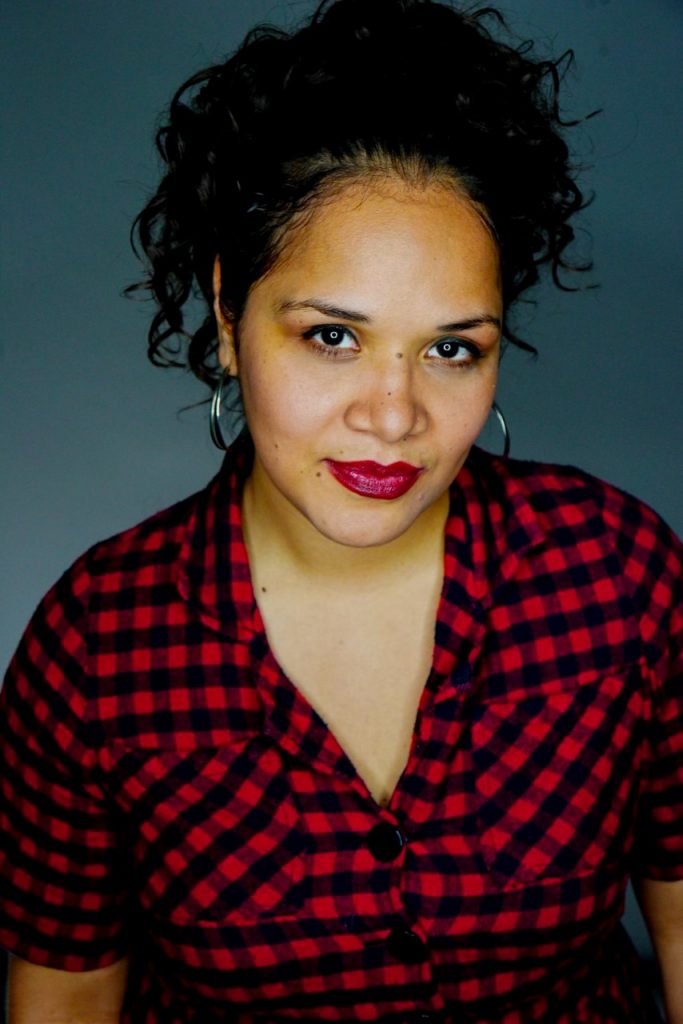 A woman with curly hair wears a red and black checkered shirt, smiling confidently against a gray background.