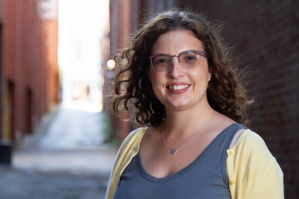 A smiling woman with curly hair stands in an alley, wearing glasses and a gray top with a yellow cardigan.